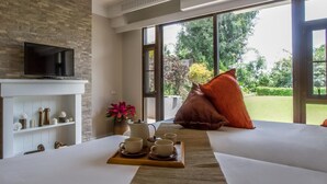 A hotel room with a television on a stone wall, a bed with a view of greenery, and a breakfast tray with cups and a flower vase. - Doi Tung Lodge (Mae Fa Luang)