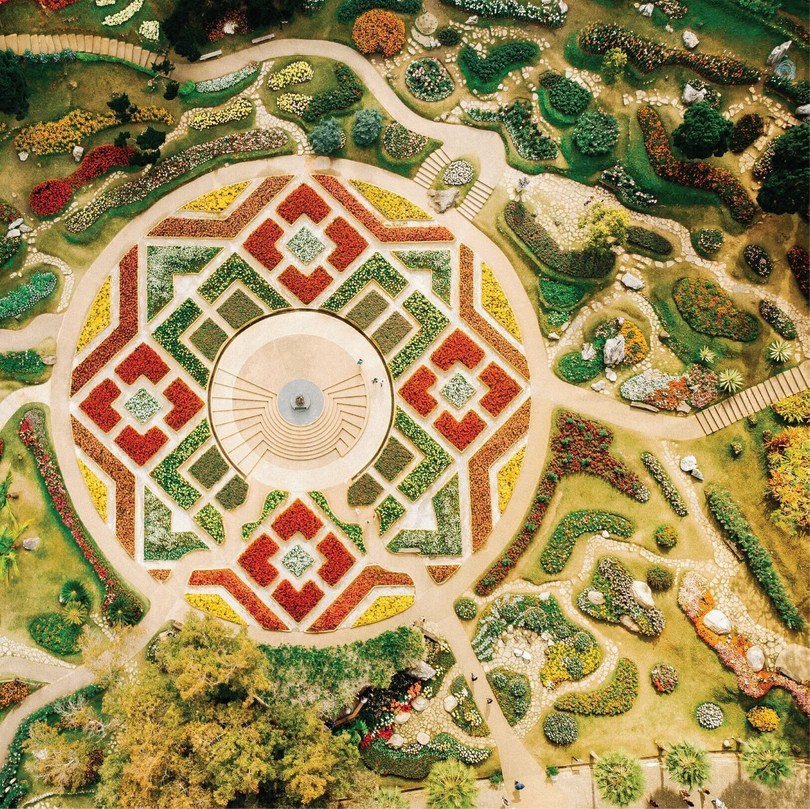 Ein kreisförmiger Garten mit einem zentralen Brunnen, umgeben von unterschiedlich gestalteten Blumenbeeten und Wegen.