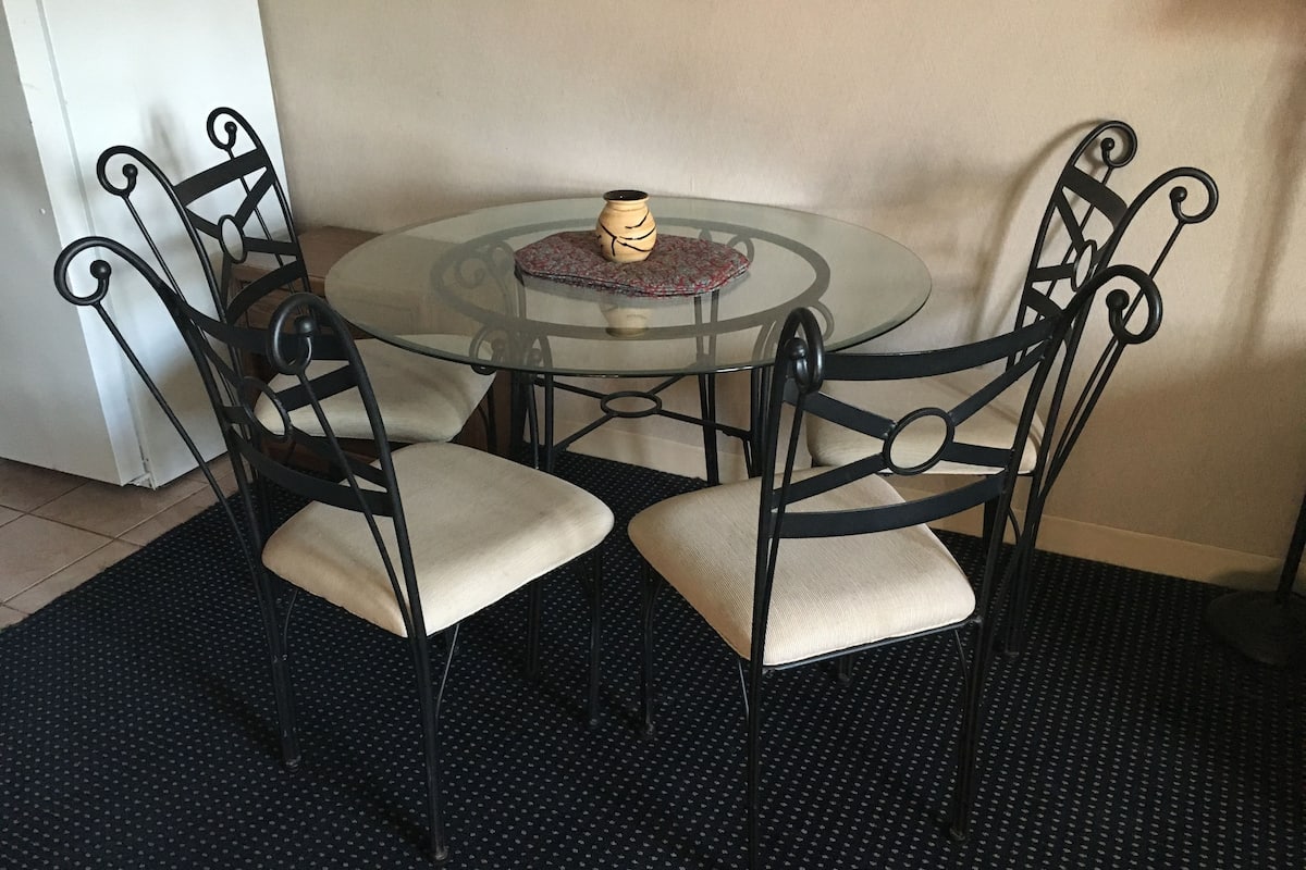A round glass table with four black metal chairs, a small decorative item on the table, and a black textured rug on the floor.