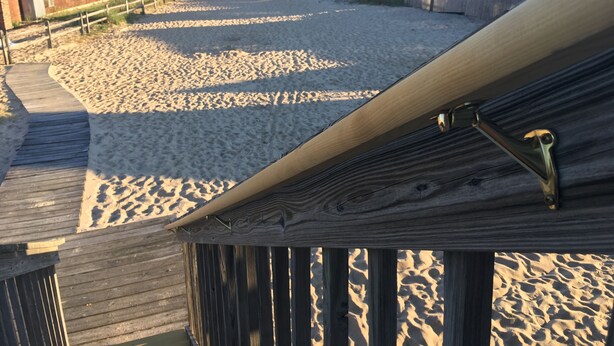 A beach view from a wooden deck with a sandy beach, a wooden fence, and a building in the background. - Beachfront White Sands (North Truro)