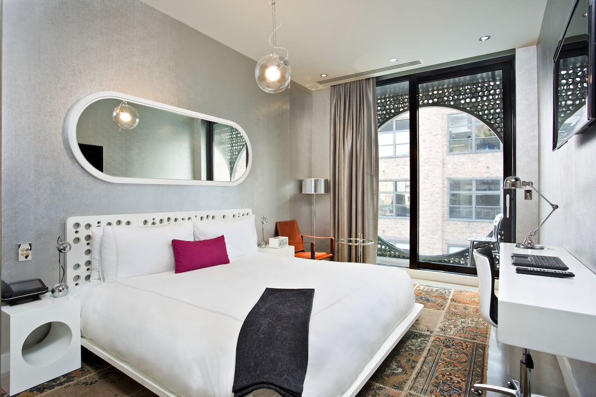 A modern hotel room with a large bed, a desk, and a view of the city through a large window.