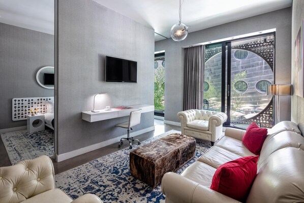 A modern living room with a flat-screen TV, a white desk, a leather sofa, and a large window with a view of greenery.