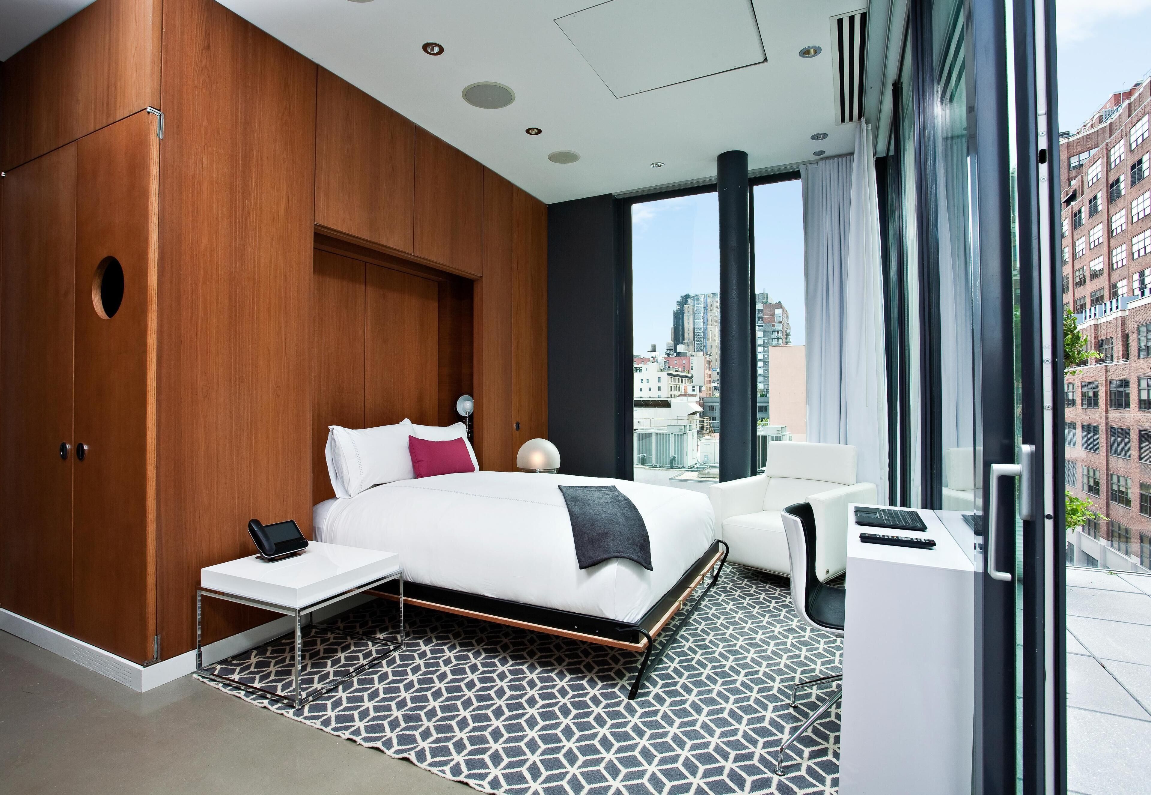 A modern hotel room with a large bed, a desk, a chair, and a view of the city through large windows.
