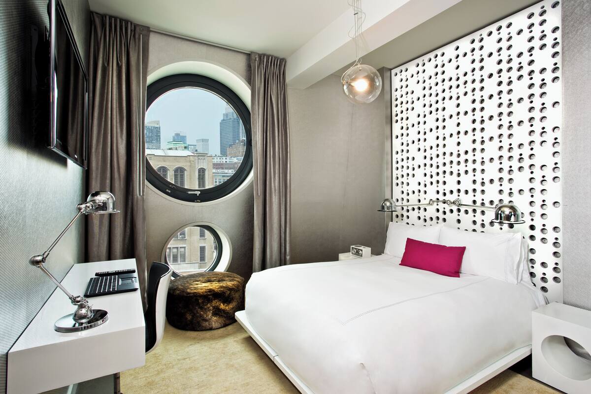 A modern hotel room with a round window, a desk with a lamp, a bed with white bedding and a pink pillow, and a decorative wall panel.