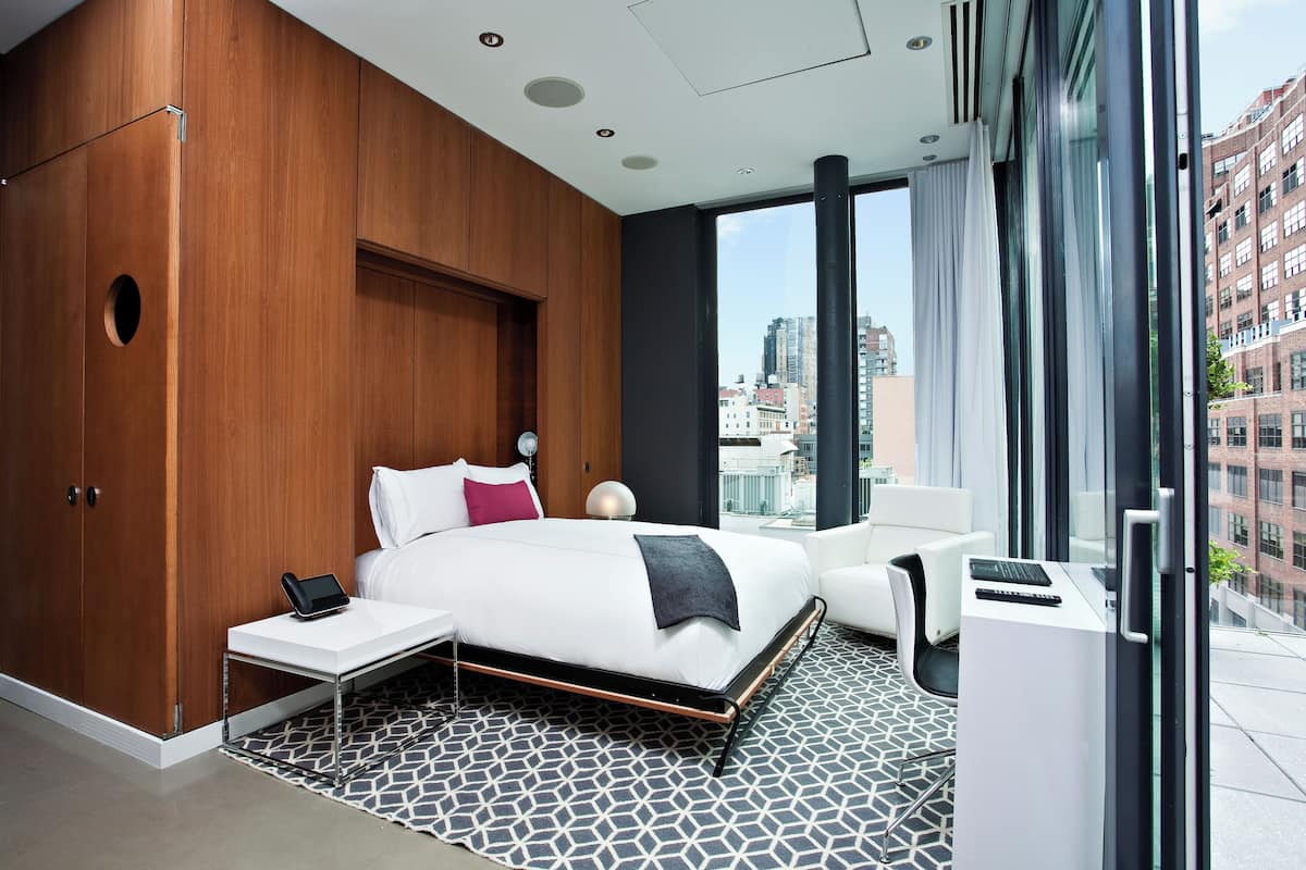 A modern hotel room with a large bed, a bedside table with a tablet, a small desk with a laptop, a large window with curtains, and a view of the cityscape.