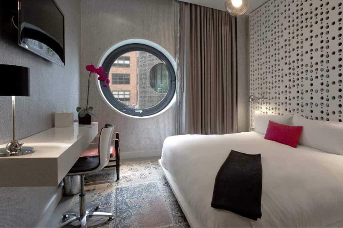 A hotel room with a large bed, a round window, a desk with a chair, and a modern wall design.
