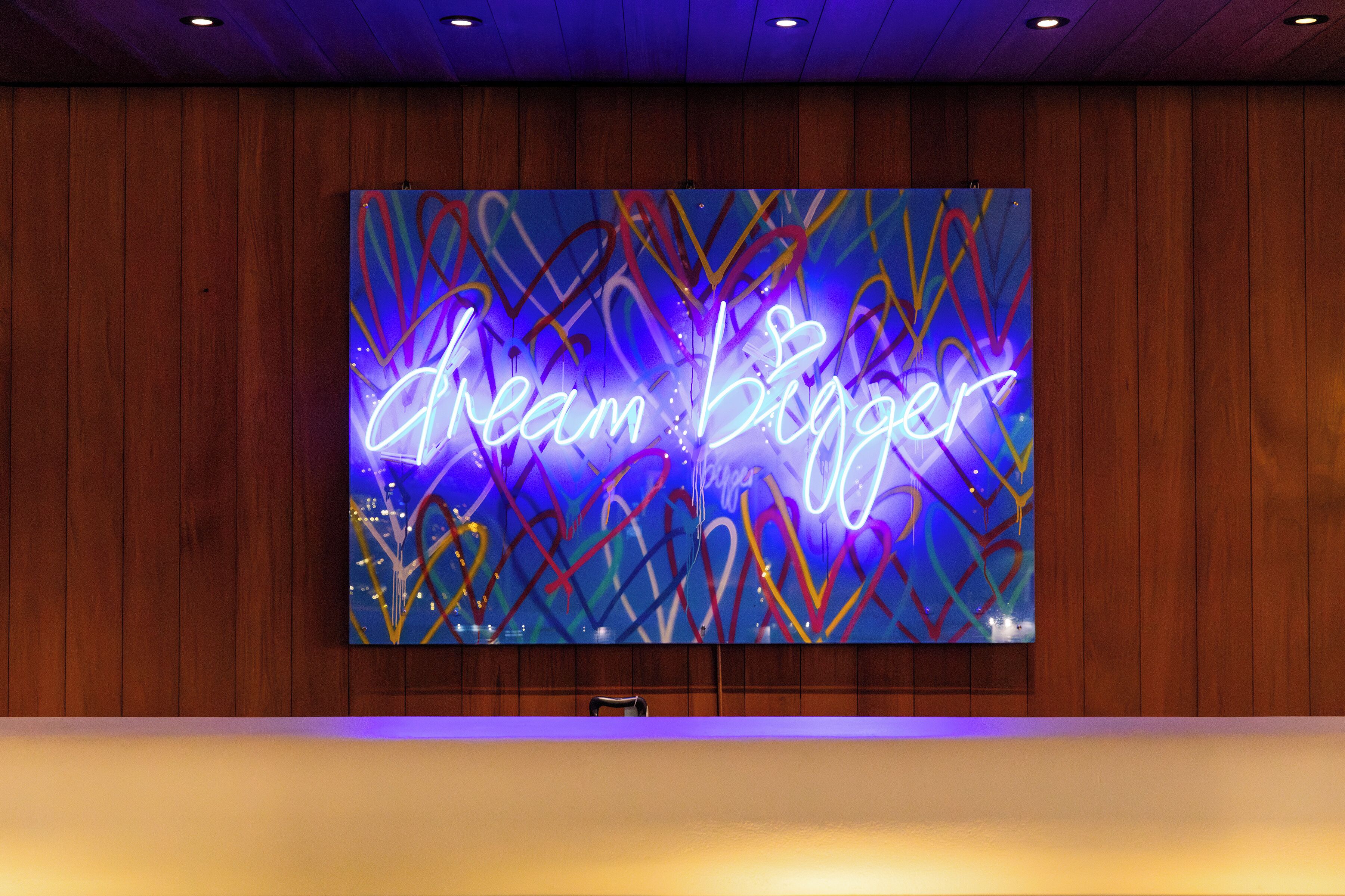 A large neon sign with the text "Dream Bigger" on a wall in a room with wooden paneling.