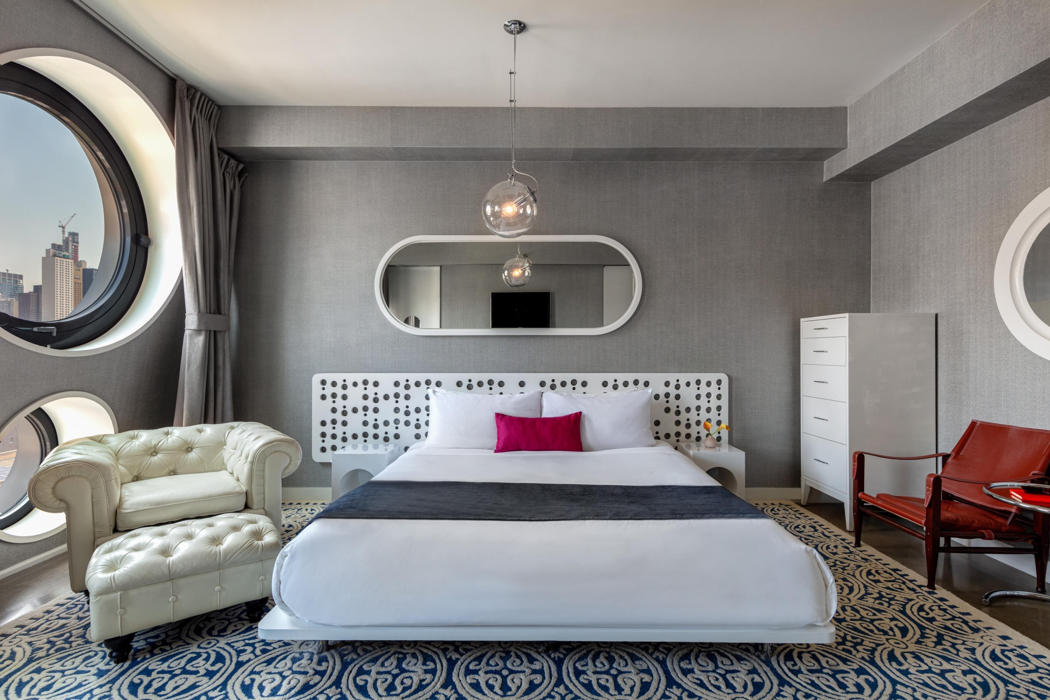 Habitación de hotel con una cama grande, un espejo redondo, vista a la ciudad, un aparador blanco, una silla roja y un tapete estampado.