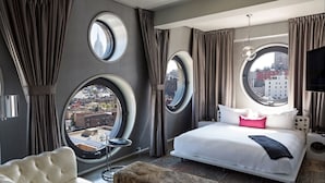 A modern hotel room with a large bed, a round window, and a view of the cityscape.