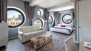 A modern hotel room with a large bed, a sofa, a coffee table, and a view of the cityscape through circular windows.