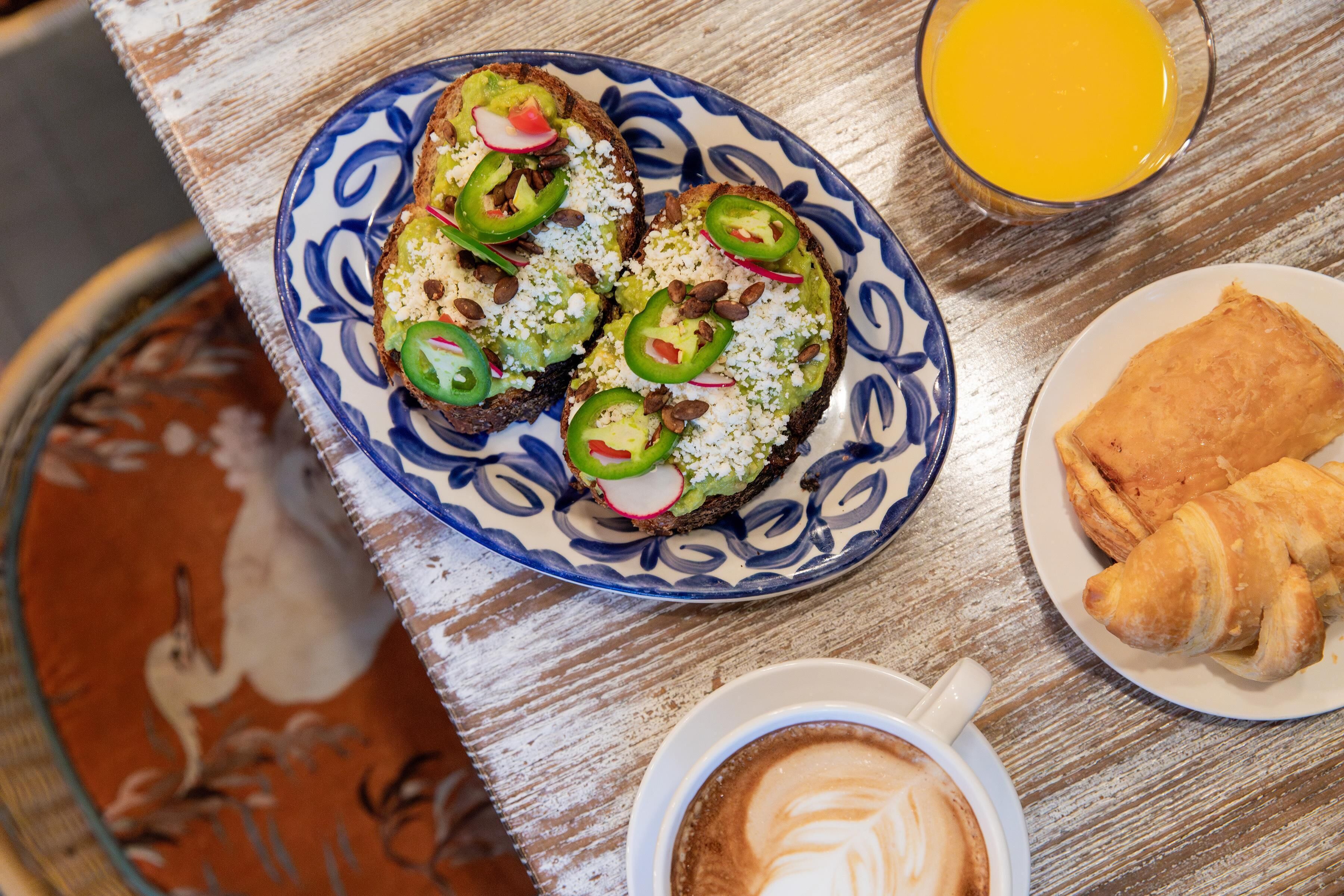 Une table dressée pour le petit-déjeuner avec des toasts garnis d’avocat, de radis et de piments jalapeño, un verre de jus d’orange, des croissants et une tasse de café décorée avec un motif en latte art.