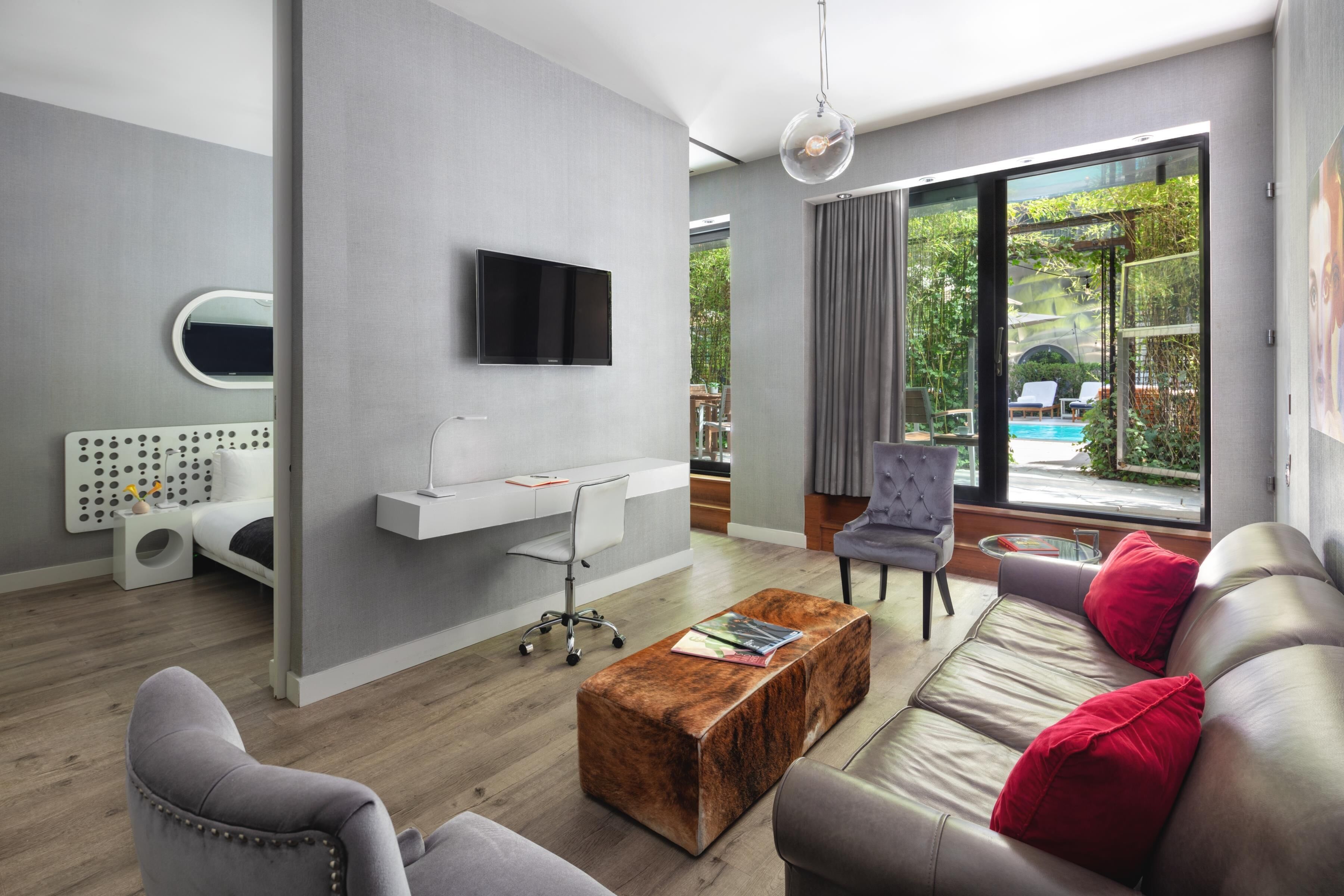 Una sala de estar moderna con un televisor de pantalla plana montado en la pared, una zona de estar con un sofá y sillones, una mesa de centro, y vista a una piscina a través de amplios ventanales.