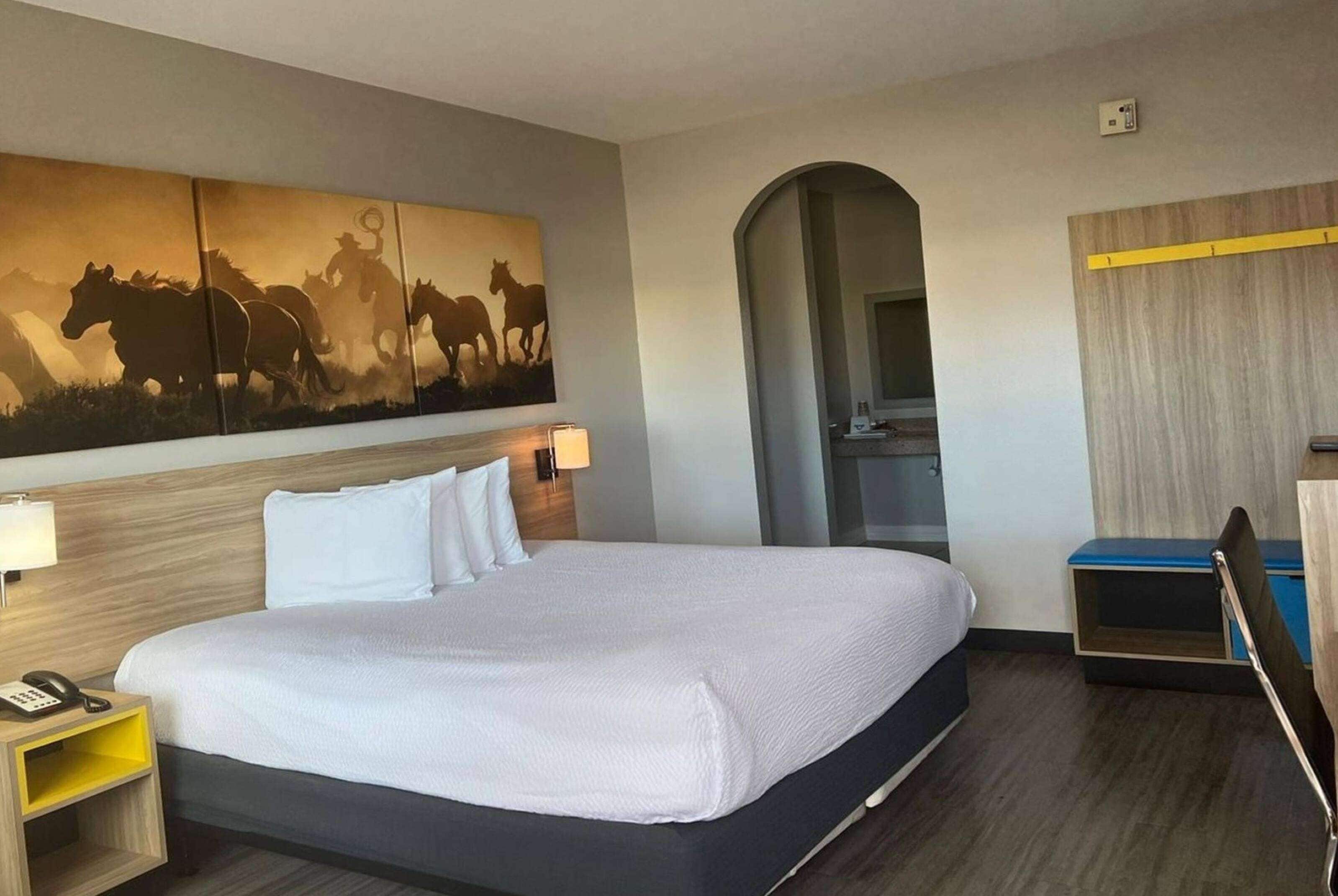 A hotel room with a large bed, a wooden headboard, a nightstand with a phone, a wall-mounted lamp, and a painting of horses.