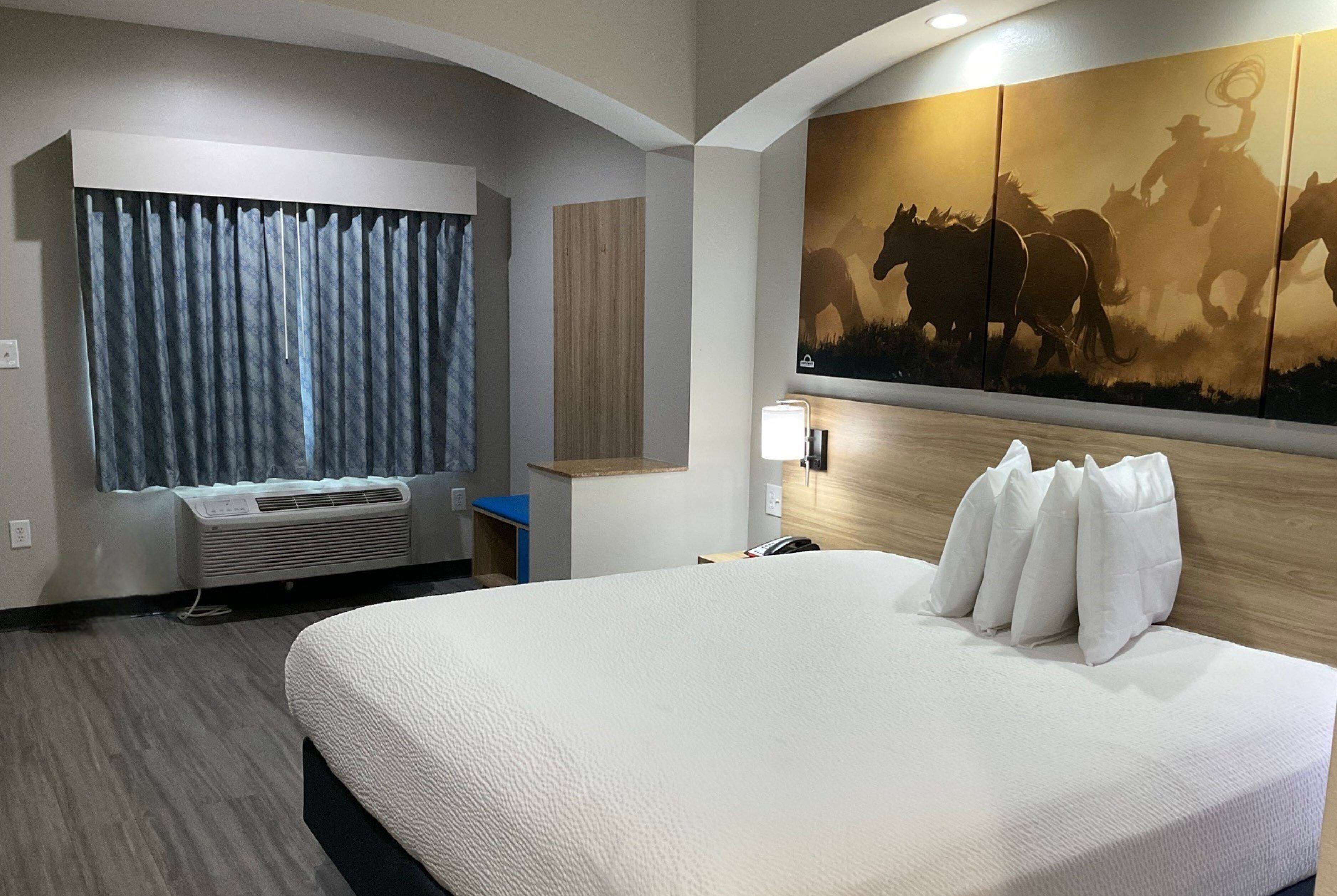Habitación de hotel con una cama grande, una obra de arte con vaqueros a caballo, un escritorio con lámpara y una ventana con cortinas.
