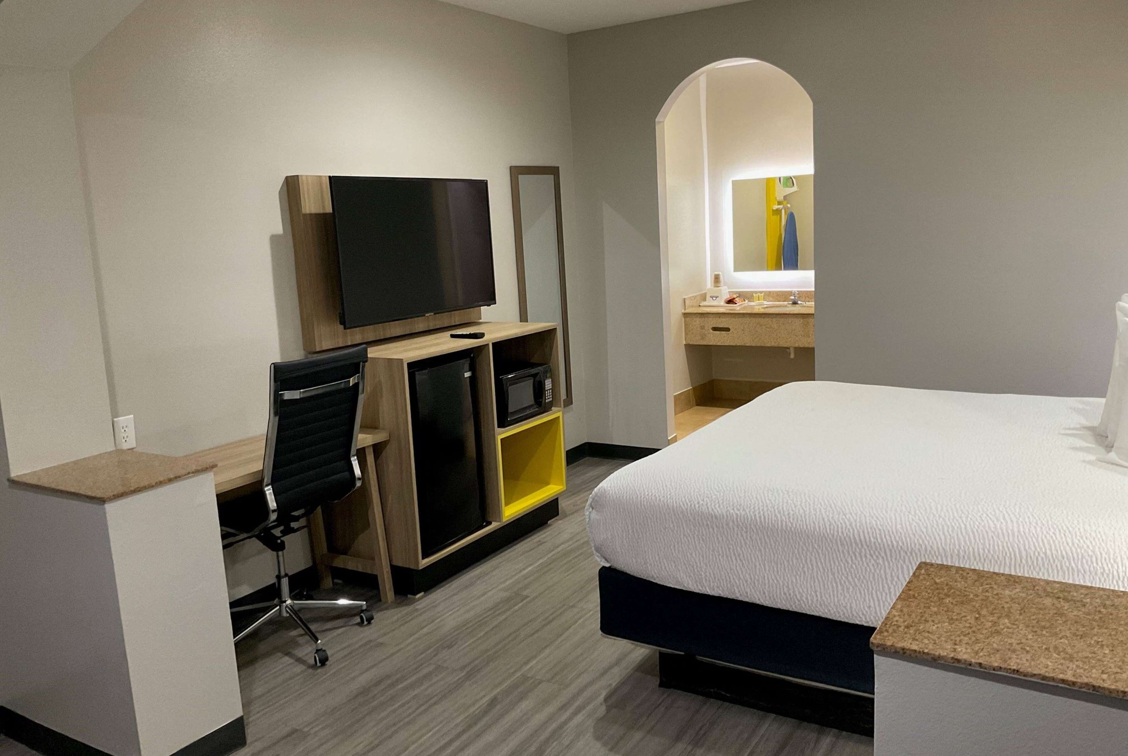 A hotel room with a bed, a desk with a computer, a TV, a microwave, and a bathroom with a mirror and sink.