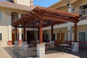 Un patio techado al aire libre con mesas y bancos de madera, una escalera y un edificio con varias puertas y ventanas.