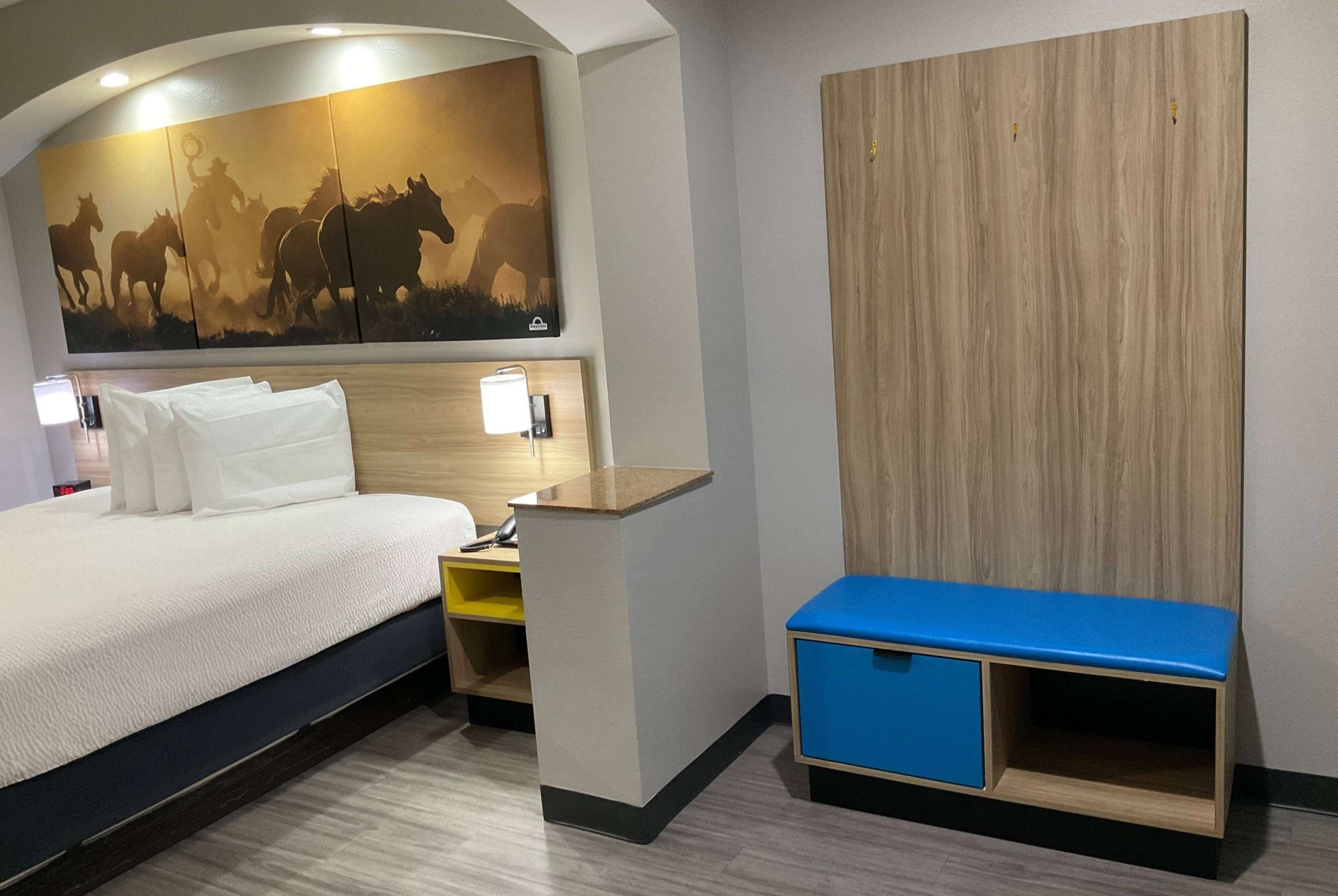 A hotel room with a bed, a desk, a blue bench, a wall-mounted artwork of horses, and a wooden panel wall.