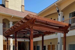 Una estructura de pérgola de madera con ventilador de techo, sostenida por columnas, frente a un edificio con varios balcones y puertas.