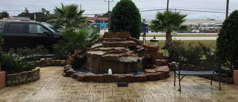 Una fuente de piedra con agua que cae, rodeada de plantas en macetas y un banco.