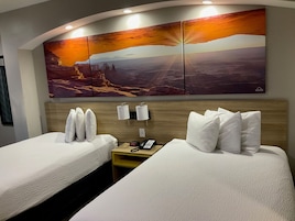 Una habitación de hotel con dos camas, un cabecero de madera, una mesita de noche con un teléfono y un cuadro grande en la pared que muestra un paisaje de atardecer.