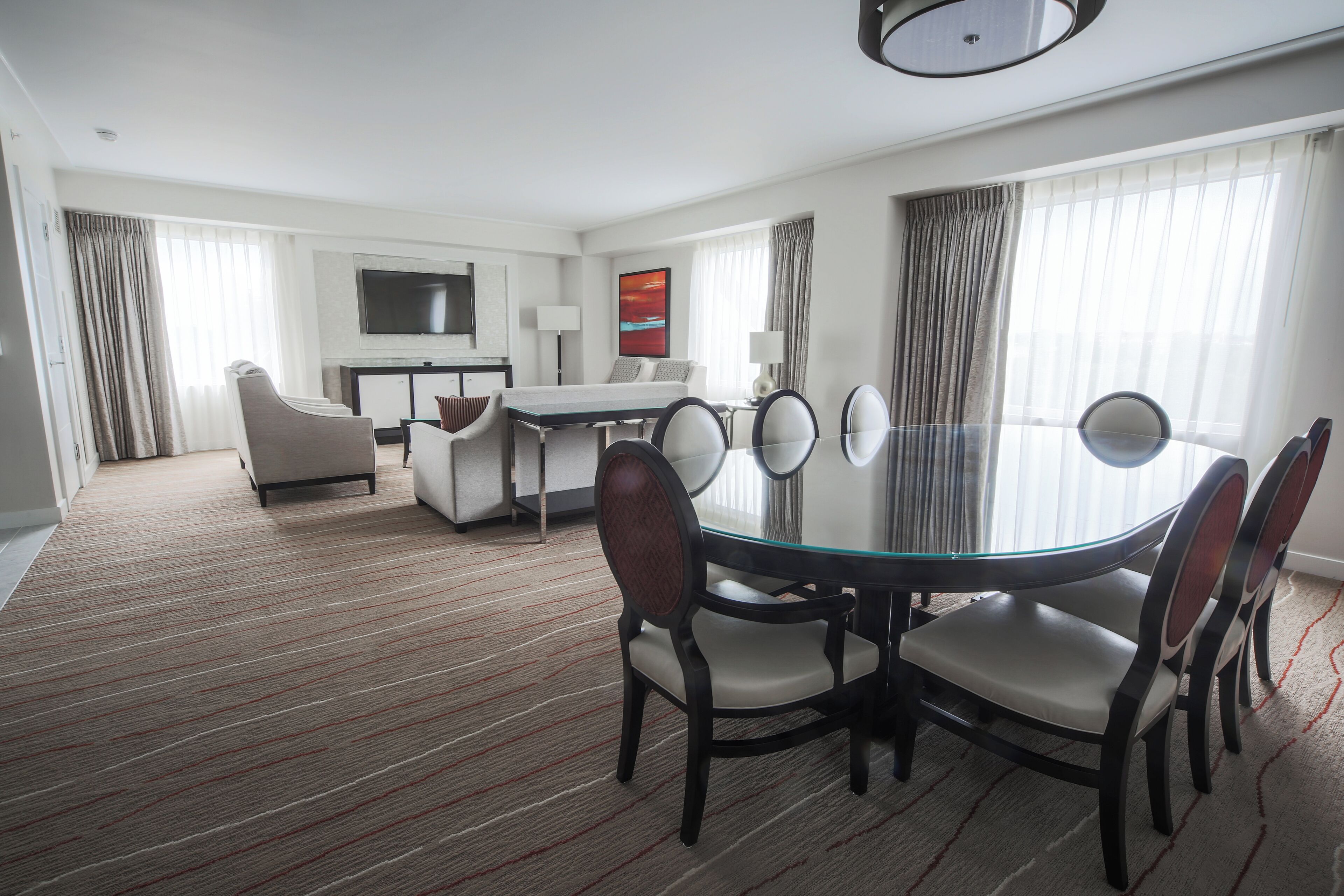 Habitación de hotel con zona de comedor, sofá, televisor y un amplio ventanal con cortinas.