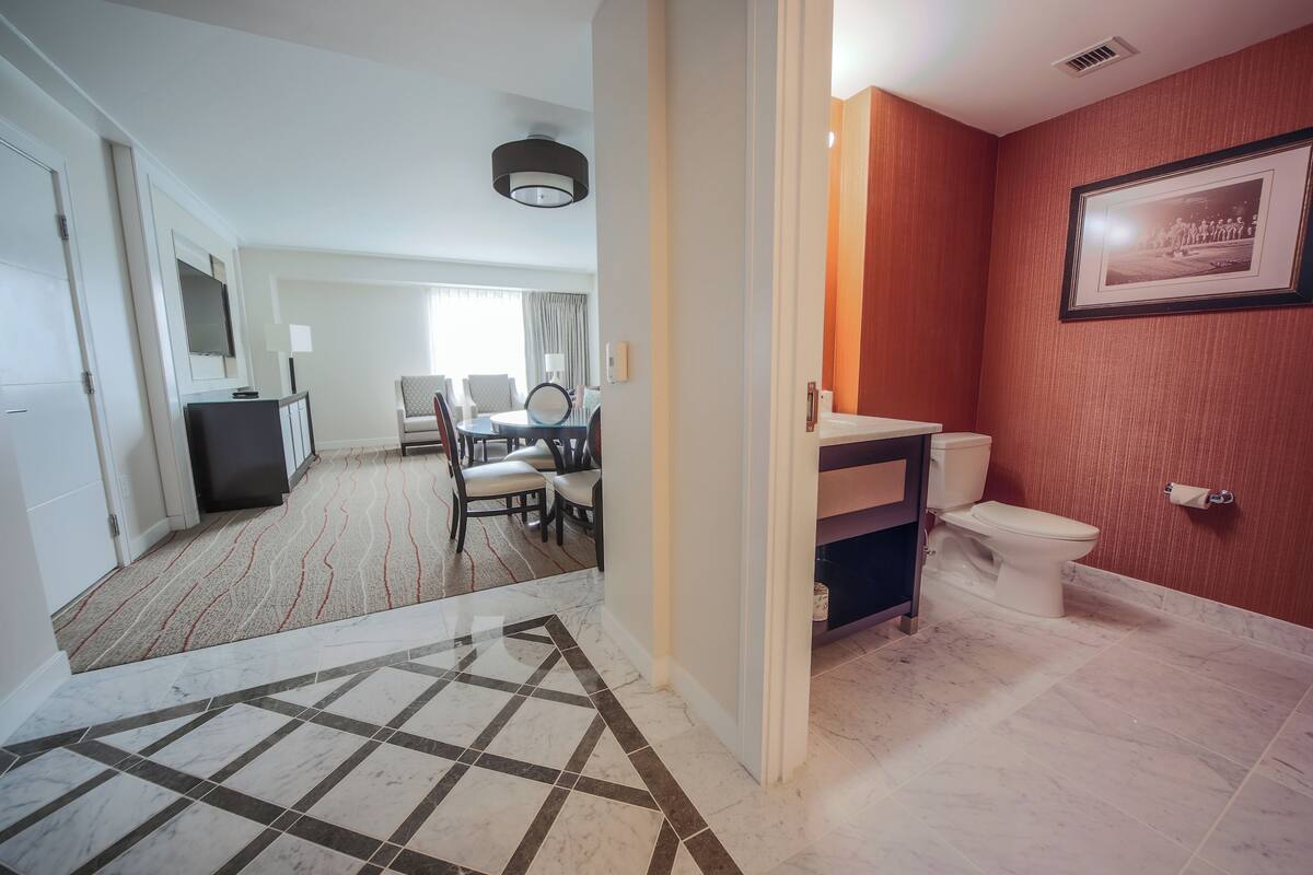 Habitación de hotel con comedor, baño con inodoro y lavamanos, y pasillo con suelo estampado.