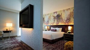 Una habitación de hotel con cama, mesita de noche, lámpara, televisor montado en la pared y alfombra estampada.