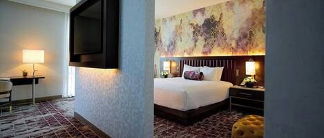 A hotel room with a bed, bedside table, lamp, TV mounted on the wall, and a patterned carpet.