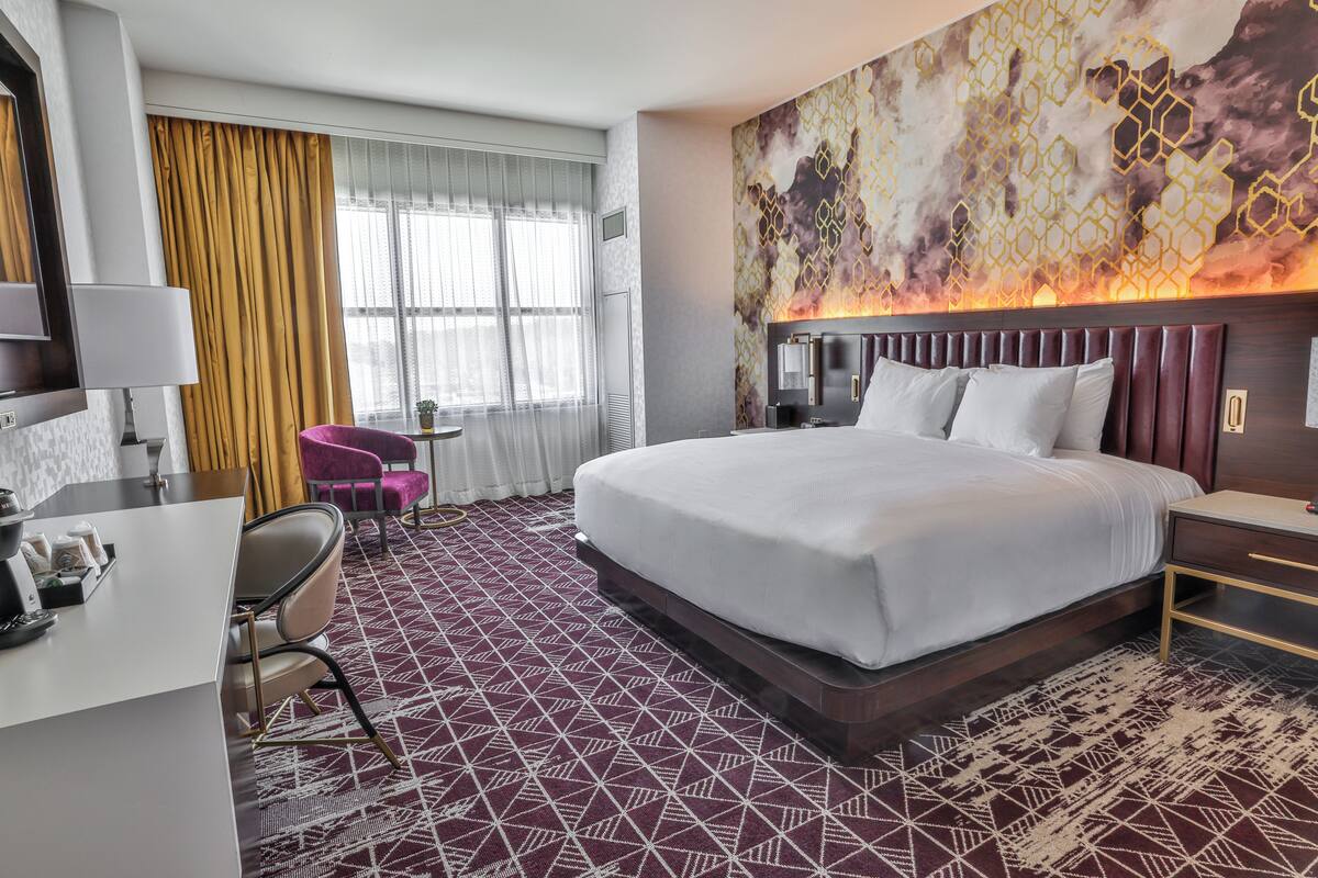 Una habitación de hotel con una cama grande, un escritorio con una silla, una mesita y un ventanal con cortinas.