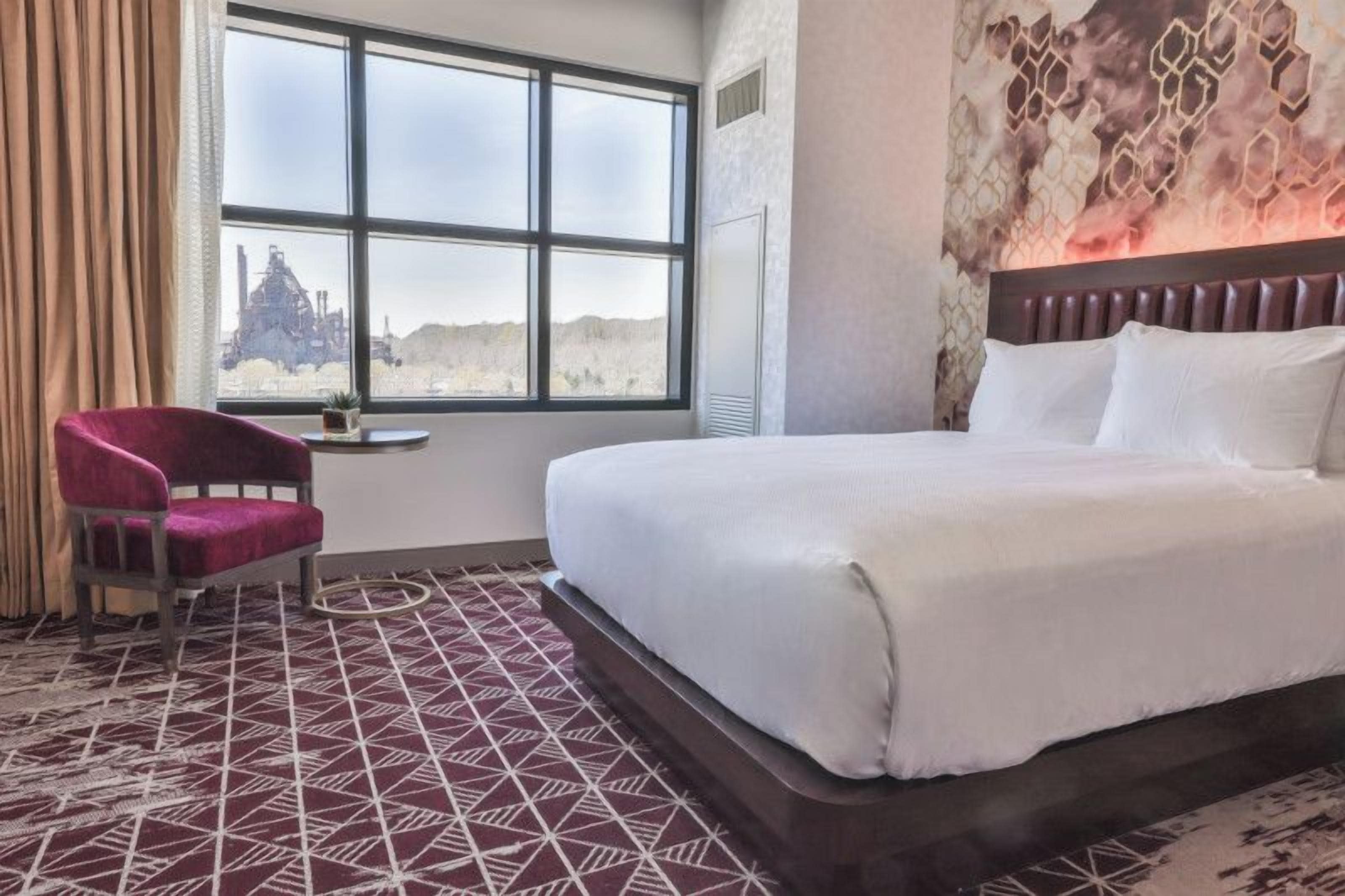 A hotel room with a large bed, a chair, a small table, and a window with a view of a distant landscape.