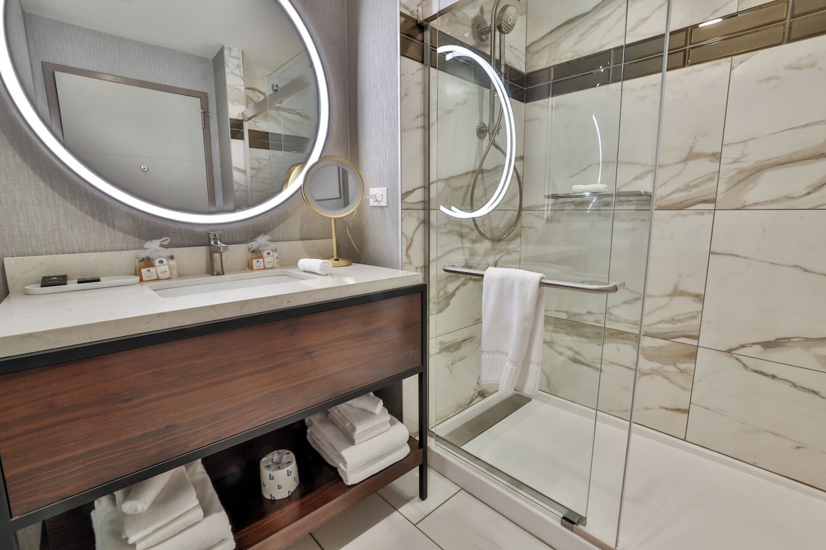 A modern bathroom with a marble-tiled shower, a round mirror, and a vanity with a dark wood finish.