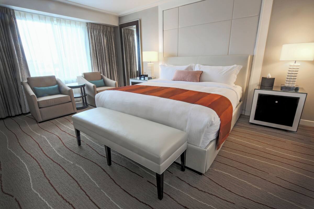 Habitación de hotel con una cama grande, un banco, dos sillones, una mesita pequeña y una mesita de noche con lámpara.