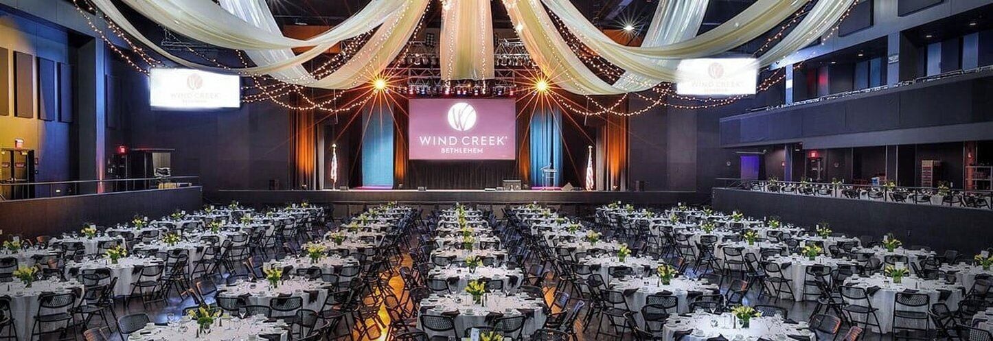 Un salón de banquetes con mesas dispuestas para un evento formal, un escenario con una pantalla grande y cortinas decorativas.