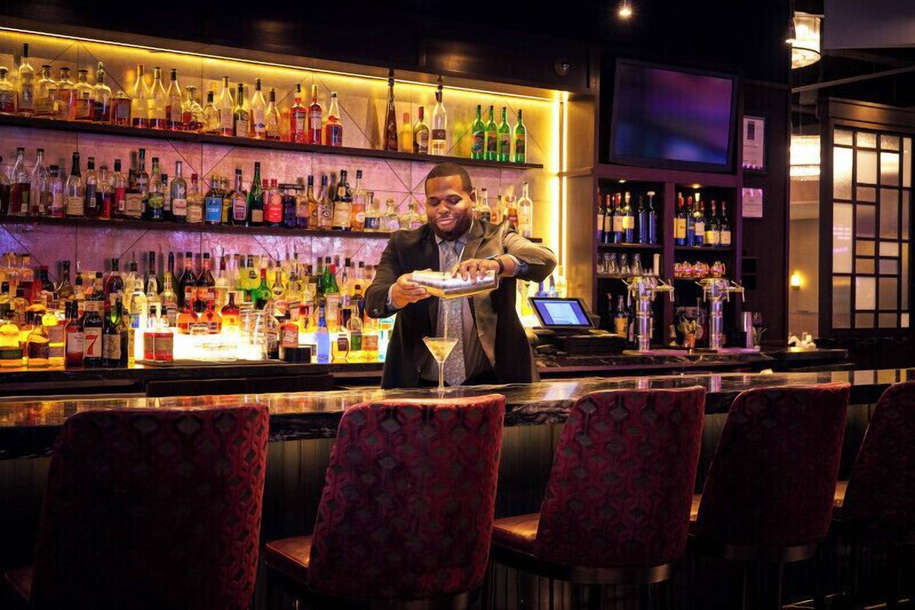 A bar area with a bartender preparing a drink, shelves of liquor bottles, and bar stools.