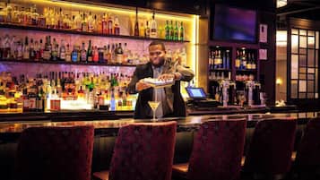 A bar area with a bartender preparing a drink, shelves of liquor bottles, and bar stools.