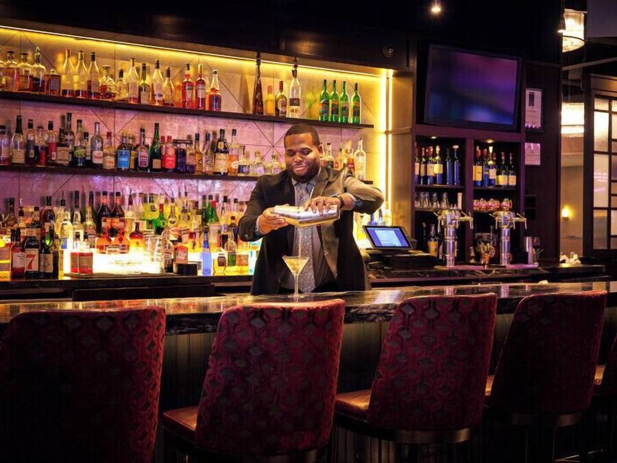 A bar area with a bartender preparing a drink, shelves of liquor bottles, and bar stools.