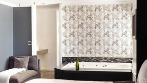 Ein modernes Schlafzimmer mit einem großen Bett, einer Couch, einem kleinen Tisch mit Flasche und Glas sowie einer Wand mit einem sich wiederholenden Muster.