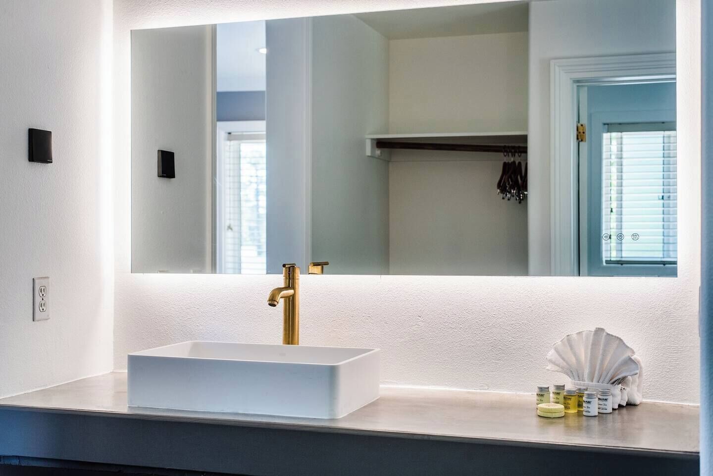 A bathroom with a white sink, gold faucet, and a large mirror. There are toiletries on the counter and a door with a window in the background.