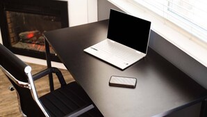 Ein Laptop auf dunklem Tisch, daneben ein Smartphone, ein schwarzer Stuhl und im Hintergrund ein Kamin.