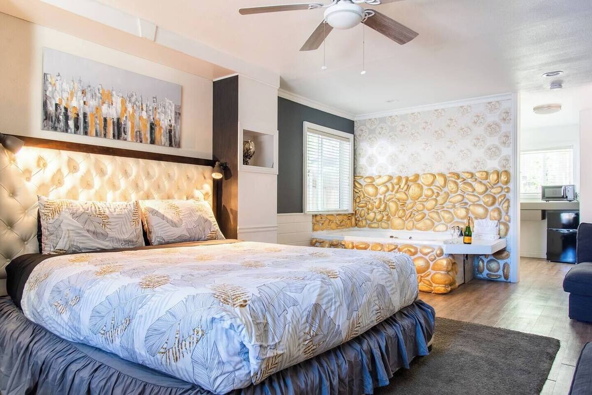 Une chambre avec un grand lit, un ventilateur de plafond, une salle de bain avec un mur en pierre et un coin cuisine.