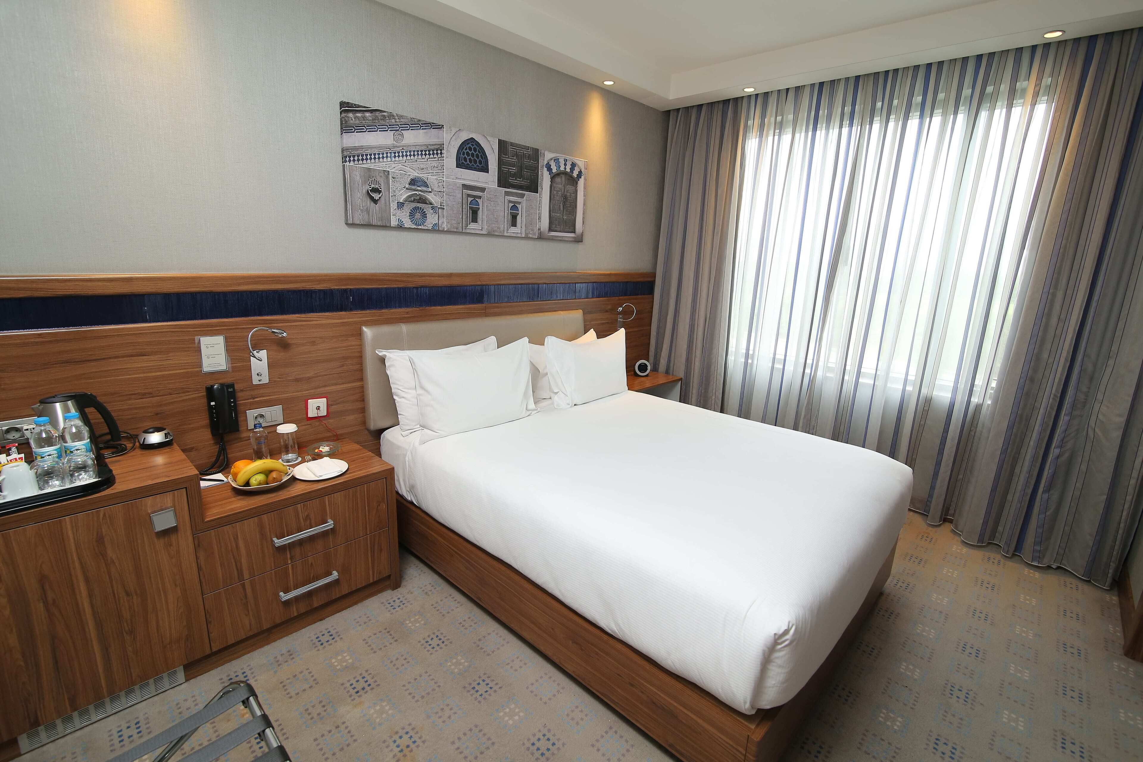 Une chambre d’hôtel avec un grand lit, une tête de lit en bois, une table de chevet avec une lampe, un petit bureau avec un téléphone et une machine à café, et un mur orné de carreaux décoratifs.