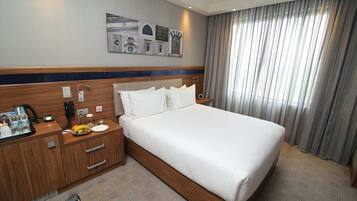 Une chambre d’hôtel avec un grand lit, une tête de lit en bois, une table de chevet avec une lampe, un petit bureau avec un téléphone et une machine à café, et un mur orné de carreaux décoratifs.