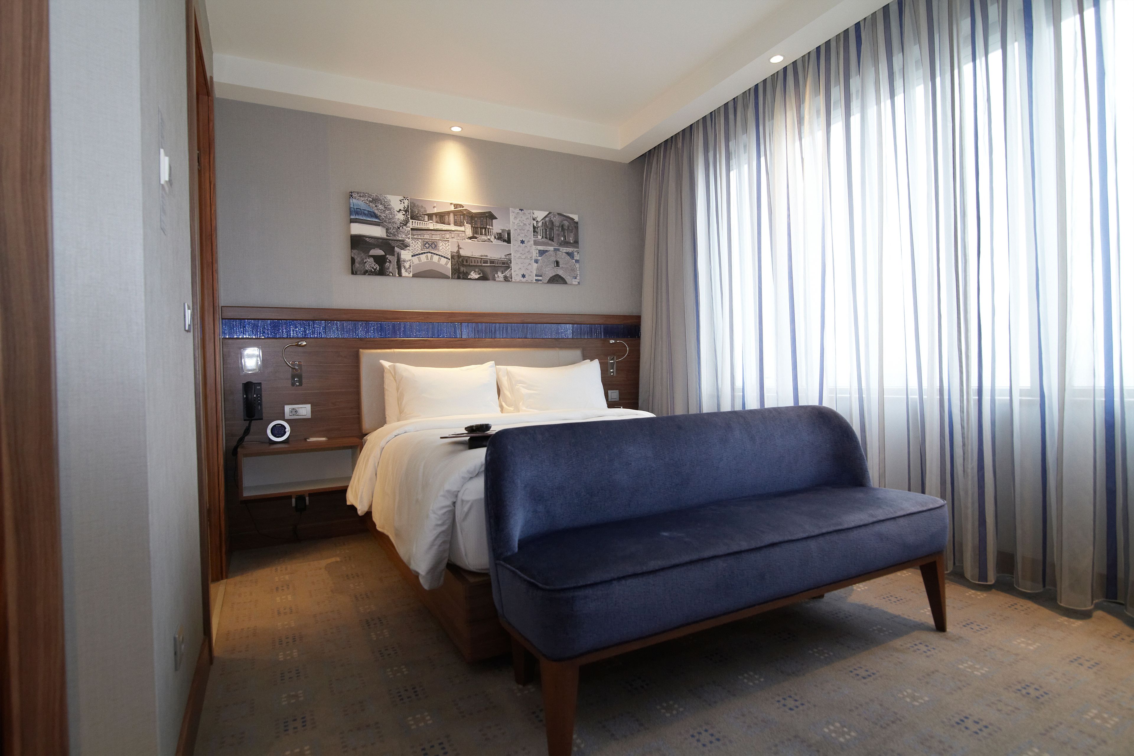 A hotel room with a bed, a blue sofa, a nightstand, a wall-mounted artwork, and a large window with curtains.