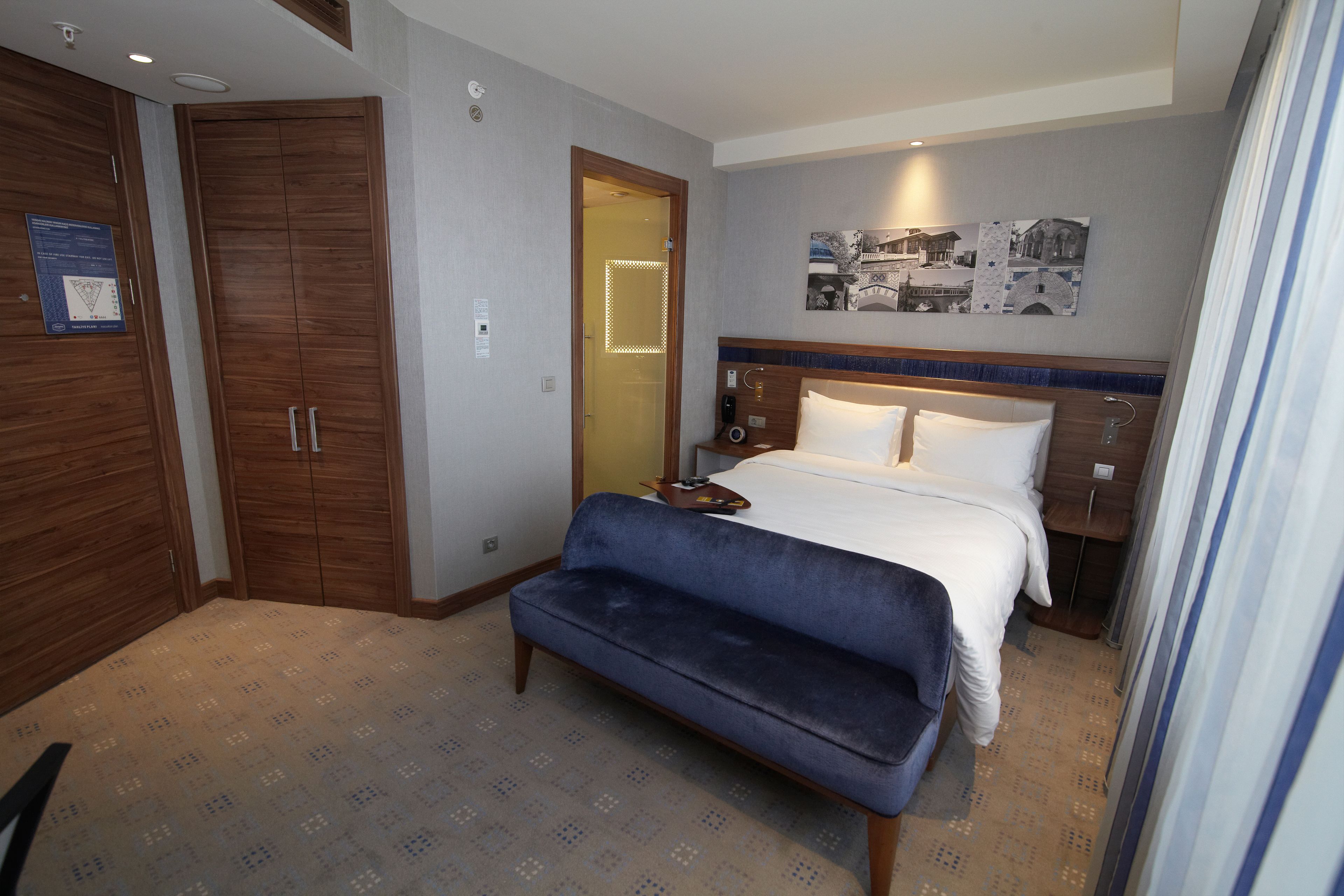 Habitación de hotel con una cama grande, un banco, un armario y un baño visible a través de una puerta abierta.