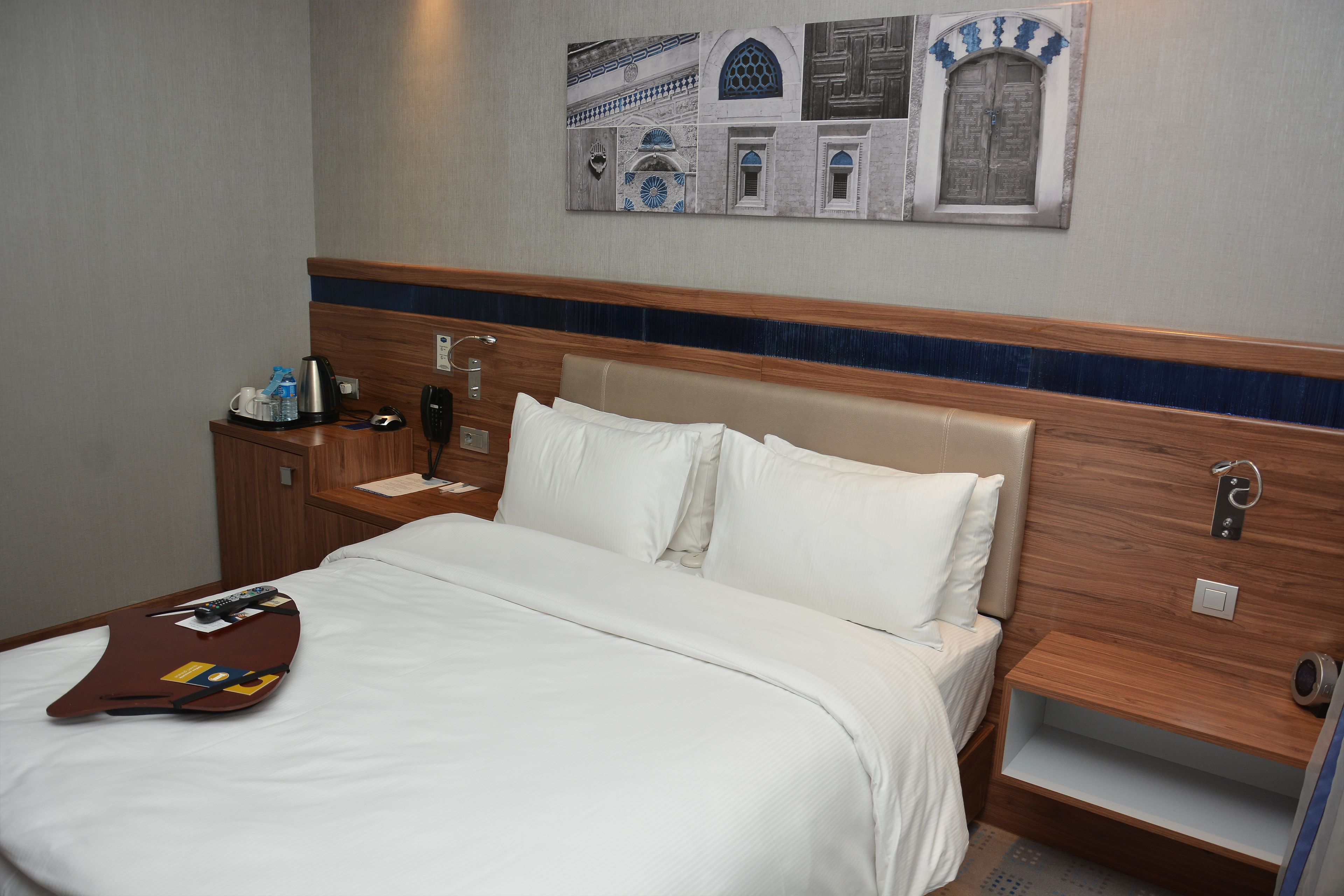 Una habitación de hotel con una cama grande, un cabecero de madera, una mesita de noche con teléfono y lámpara, y una pared con un panel decorativo.