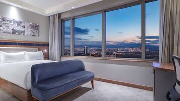 Habitación de hotel con cama, sofá azul, escritorio con silla y un ventanal que ofrece vistas de la ciudad.