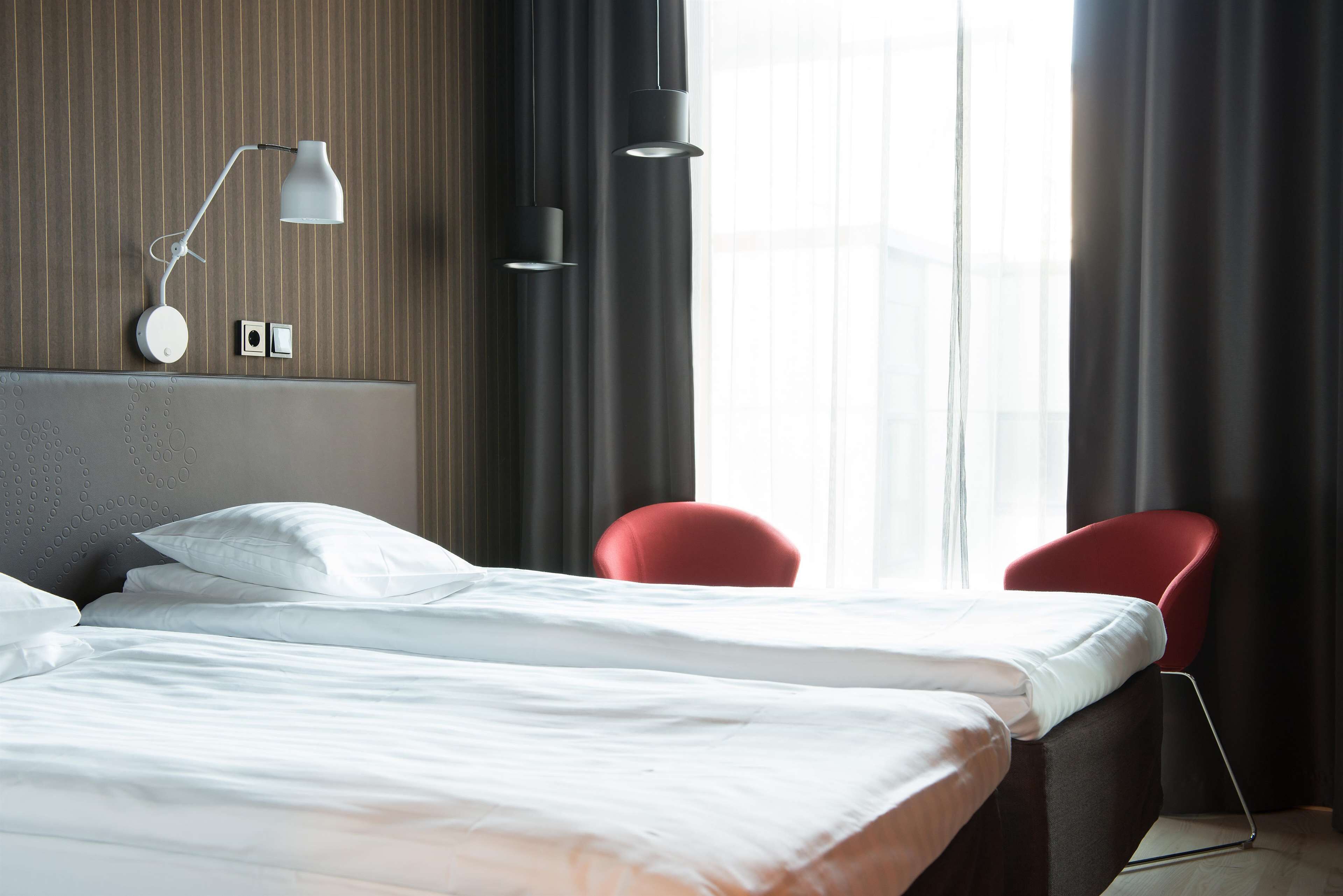 A modern hotel room with a large bed, two red chairs, a wall-mounted lamp, and a window with curtains.