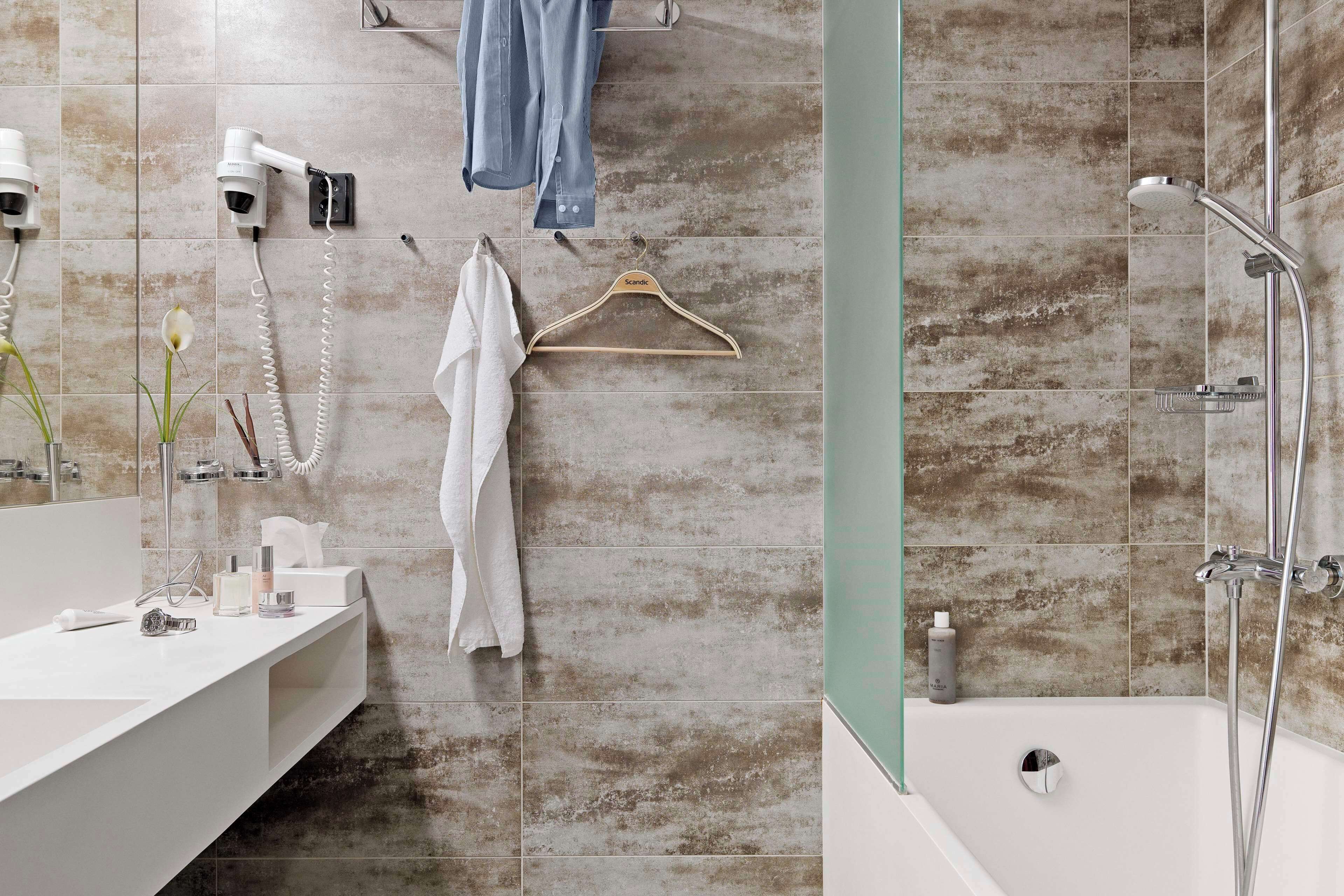 Un baño con un lavamanos blanco, un perchero con una toalla blanca, un secador de pelo y una ducha con cabezal de ducha de mano.