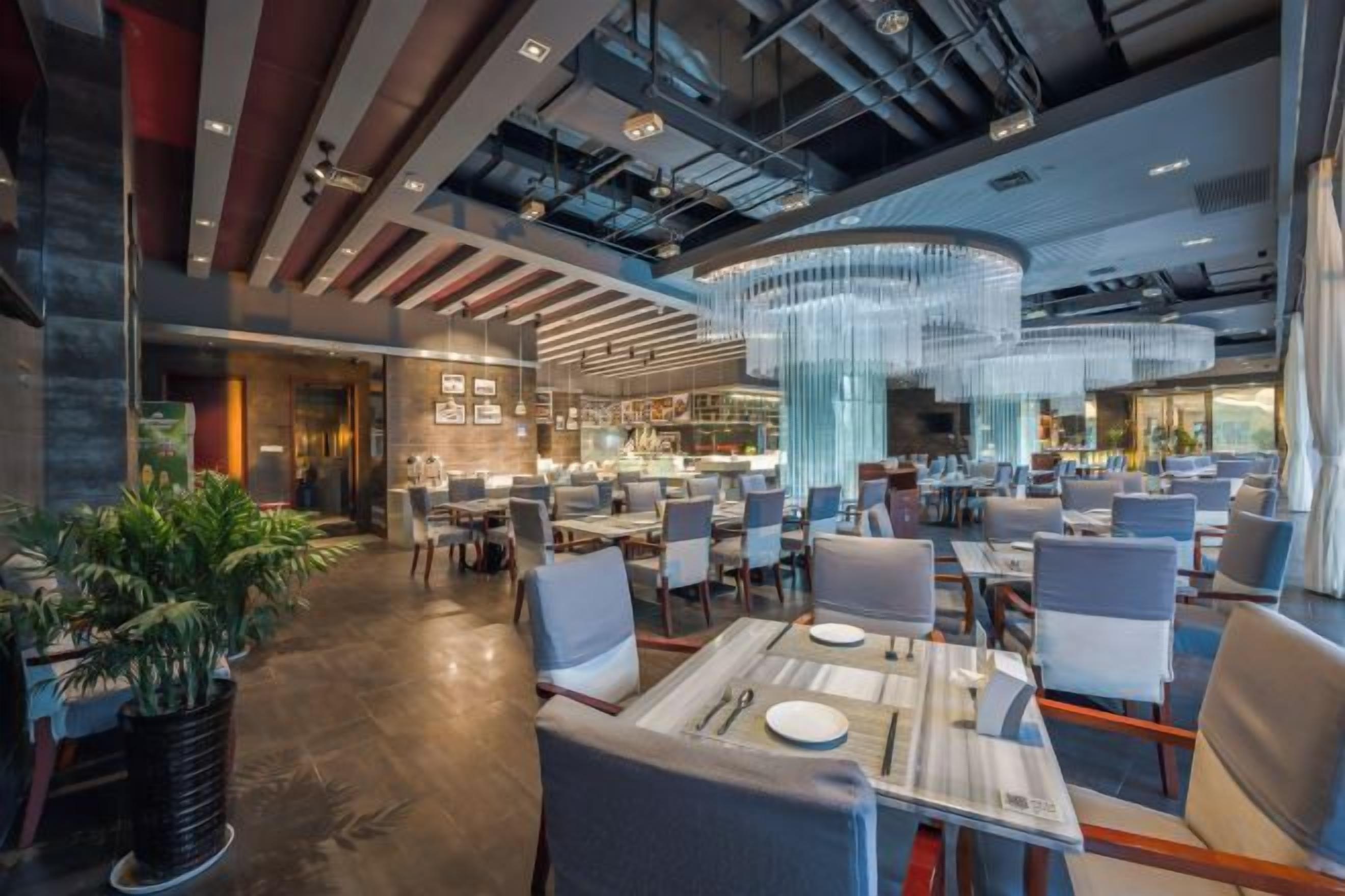 Un restaurante moderno con techos altos, conductos expuestos y una lámpara central. Las mesas están ordenadas con sillas, y hay plantas en macetas en el suelo.