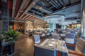 A modern restaurant with high ceilings, exposed ductwork, and a central chandelier. Tables are neatly arranged with chairs, and there are potted plants on the floor. - Harbour Oriental Hotel（Beilun Changjiang Road Store） (Ningbo)